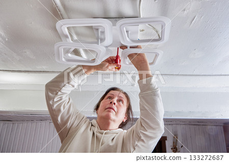 Positioning geometric LED fixture on ceiling involves woman using screwdriver for adjustment, modern lighting shape and tool grip are clearly observed. Positioning geometric LED fixture on ceiling involves woman using screwdriver for adjustment, modern lighting shape and tool grip are clearly observed. 133272687