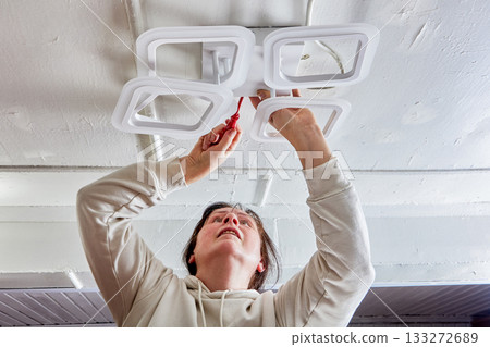 Mounting LED chandelier on ceiling in rural house is carried out by woman, modular light fixture and hand tool are visible, installation sequence underway. 133272689