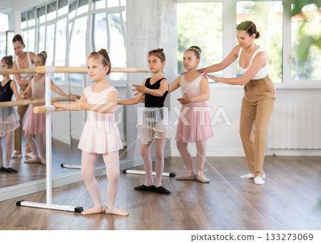 Teacher conducts class with children girls in ballet studio, observes doing grand plie exercise. 133273069