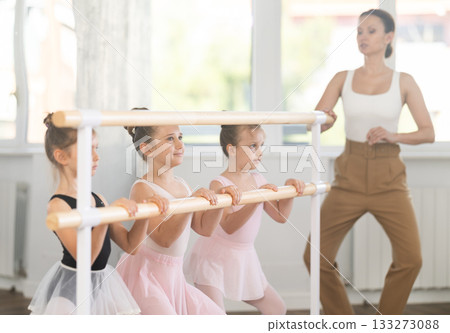Teacher conducts class with children girls in ballet studio, observes doing grand plie exercise. 133273088