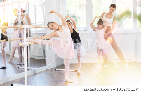 Girl ballet students prepare for classes, near ballet bare stretch their leg under teacher guidance. Girl ballet students prepare for classes, near ballet bare stretch their leg under teacher guidance. 133273117
