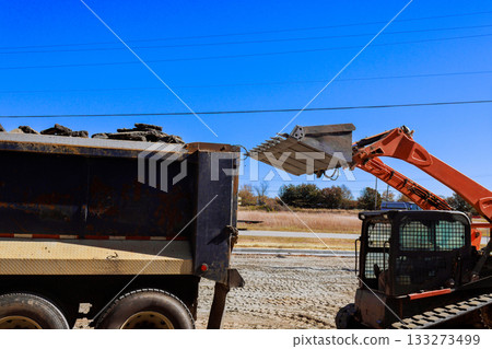 Skid steer machinery loads dirt into dump truck at construction site 133273499