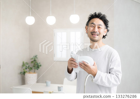 A man doing housework while wiping dishes with a smile in a bright kitchen, looking at the camera A man doing housework while wiping dishes with a smile in a bright kitchen, looking at the camera 133276286