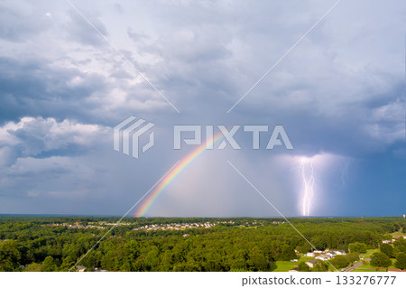 Vibrant rainbow spans sky as storm brews, lightning striking in distance over lush greenery. 133276777