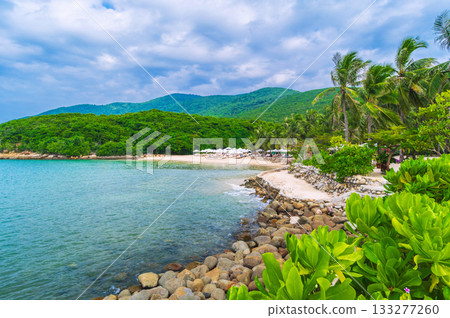 panoramic landscape with a sandy beach with sun umbrellas and sun beds under palm trees in bay by the sea on cloudy summer day at resort on a paradise island in Asia 133277260