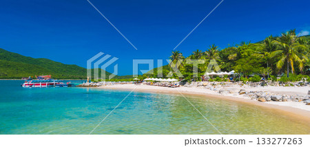 landscape panorama with white sandy beach with sun loungers and umbrellas under palm trees in bay by sea on a sunny summer day at a resort on paradise island 133277263