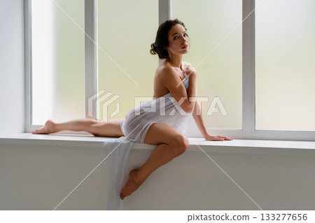 Woman in white fabric poses gracefully by large window in natural light 133277656
