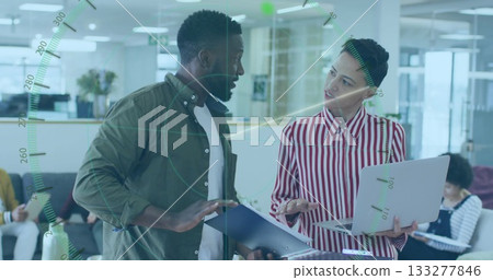 Two professionals discussing work over document folder and laptop in open plan office, with sofa 133277846