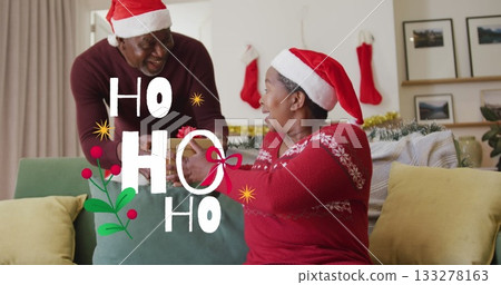 Santa-hatted man giving ribbon-tied gift to woman on sofa in living room, with stockings and tinse Santa-hatted man giving ribbon-tied gift to woman on sofa in living room, with stockings and tinse 133278163