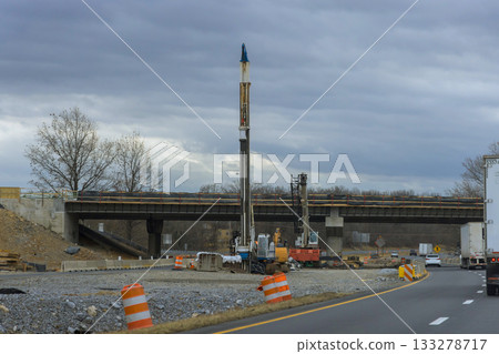 Workers are setting up drilling equipment next to highway under construction with partially built overpass overhead. Workers are setting up drilling equipment next to highway under construction with partially built overpass overhead. 133278717