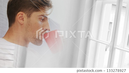 Man sipping from red ceramic mug near white-framed window in home living area, with striped cushion Man sipping from red ceramic mug near white-framed window in home living area, with striped cushion 133278927