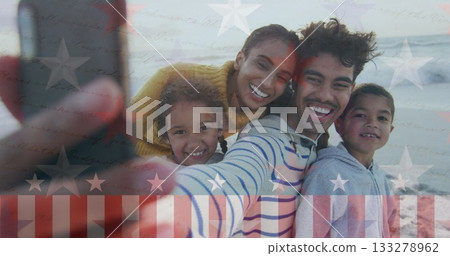 Taking family selfie on sandy ocean shoreline, showcasing smartphone with stars and stripes overlay 133278962