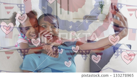 Smiling mother and daughter taking selfie on sofa at home, with smartphone digital hearts overlay 133278965