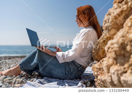 Laptop beach remote work redhead woman works on a pebble beach enjoying sunny day Laptop beach remote work redhead woman works on a pebble beach enjoying sunny day 133280449