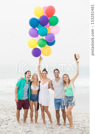 Cheerful caucasian young friends with colorful balloons standing at beach against clear sky Cheerful caucasian young friends with colorful balloons standing at beach against clear sky 133281481