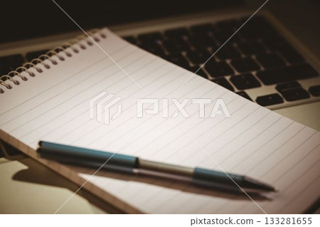 Flat design notebook with ballpoint pen is resting on laptop keyboard over desk with warm lighting 133281655