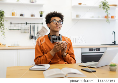 A young man in glasses holds a coffee cup while looking up thoughtfully in a bright kitchen setting. 133282464