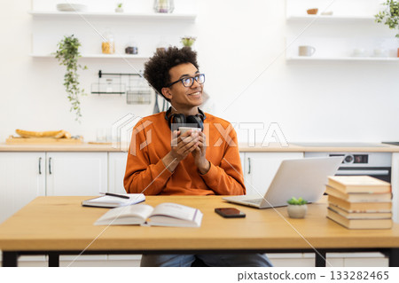 A young man with glasses smiles while holding a cup of coffee in his kitchen, with a laptop and books on the table. A young man with glasses smiles while holding a cup of coffee in his kitchen, with a laptop and books on the table. 133282465