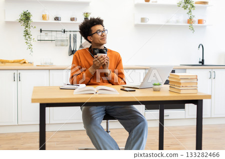 A young man with glasses enjoys a cup of coffee while working on his laptop in a bright, modern kitchen setting. A young man with glasses enjoys a cup of coffee while working on his laptop in a bright, modern kitchen setting. 133282466