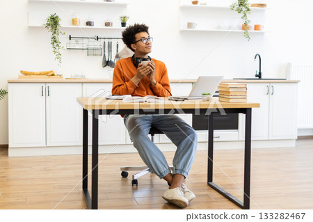 A young man with glasses enjoys a cup of coffee while working on his laptop at home in his kitchen. A young man with glasses enjoys a cup of coffee while working on his laptop at home in his kitchen. 133282467