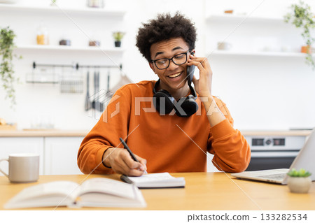 A young man with glasses smiles while talking on the phone and taking notes at his desk in a bright, modern kitchen. A young man with glasses smiles while talking on the phone and taking notes at his desk in a bright, modern kitchen. 133282534