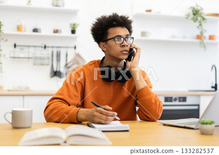 A young man with glasses is on the phone while working from home, looking concerned. A young man with glasses is on the phone while working from home, looking concerned. 133282537