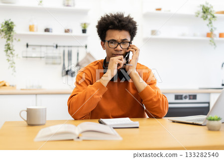 A young man with glasses is on the phone, looking concerned while sitting at a table in a kitchen setting. A young man with glasses is on the phone, looking concerned while sitting at a table in a kitchen setting. 133282540