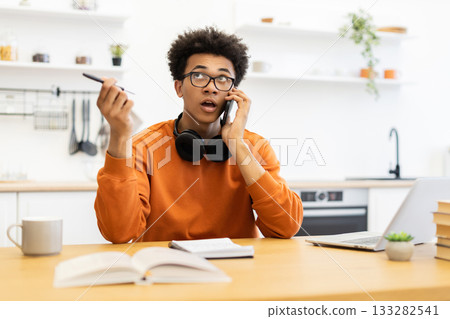 A young man with glasses is on a phone call while working at a table in a modern kitchen, holding a pen. 133282541