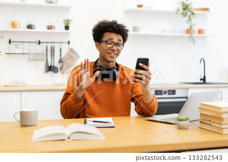 A young man with glasses waves during a video call, sitting at a table with a laptop and books in a bright kitchen setting. A young man with glasses waves during a video call, sitting at a table with a laptop and books in a bright kitchen setting. 133282543