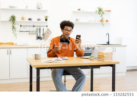 A young man with glasses is on a video call in his kitchen, looking surprised while gesturing with his hands. 133282553