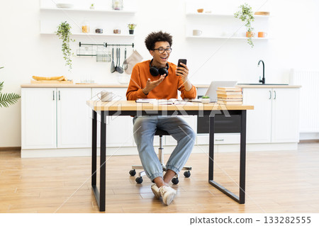 A young man with glasses is on a video call, sitting at a desk in a bright kitchen setting. 133282555