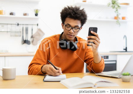 A young man with glasses is working at a table, using his phone and writing in a notebook. A young man with glasses is working at a table, using his phone and writing in a notebook. 133282558
