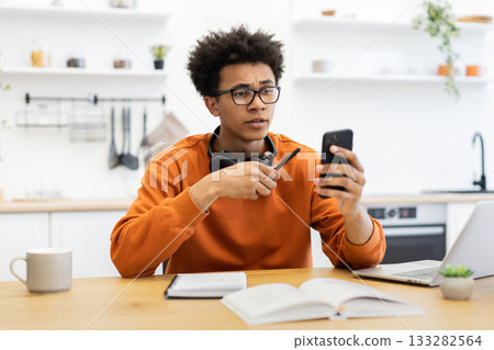 A young man with glasses looks at his phone with a concerned expression while sitting at a table in a kitchen. 133282564