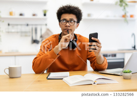 A young man in glasses contemplates while holding a phone and pen, with a book and laptop on the table. A young man in glasses contemplates while holding a phone and pen, with a book and laptop on the table. 133282566