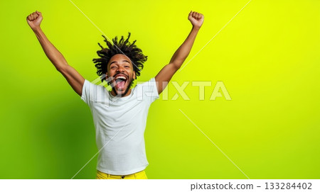 Joyful young man celebrates with raised arms against a bright green backdrop expressing happiness and excitement during a fun moment 133284402