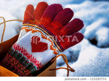 雪景背景下的紙質禮品袋,飾有紅色心形圖案,內含北歐風格的手工編織手套。 雪景背景下的紙質禮品袋,飾有紅色心形圖案,內含北歐風格的手工編織手套。 133285733