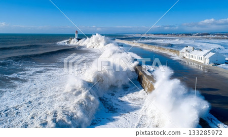 Powerful ocean waves crash against snowy coastline with lighthouse and blue sky 133285972
