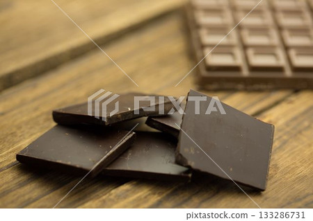 Broken dark chocolate squares are resting on flat design wooden tabletop, showing intact bar behind 133286731