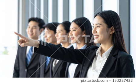 Group of professionals in business attire stand confidently by large windows, with one person pointing forward 133287422
