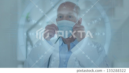 Senior doctor wearing coat adjusting surgical mask over face in hospital corridor, with tech screen Senior doctor wearing coat adjusting surgical mask over face in hospital corridor, with tech screen 133288322