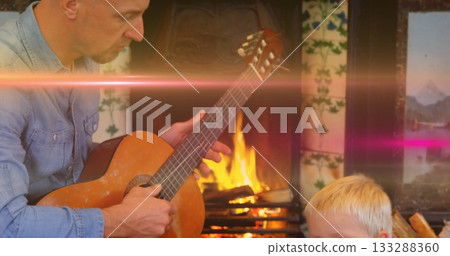 Strumming father in denim shirt teaching son guitar chords in living room tiled fireplace glowing 133288360