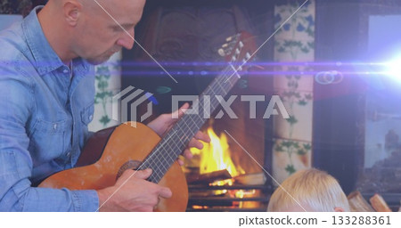 Strumming denim shirt father playing acoustic guitar beside tiled fireplace at home with lens flare 133288361