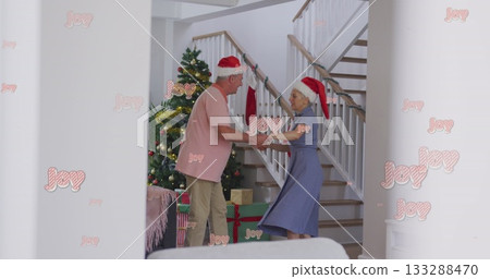 Dancing couple wearing Santa hats in hallway, with Christmas tree, presents and garlanded banister 133288470