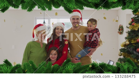 Smiling family group wearing Santa hats posing in living room, with golden snowflake confetti 133288475