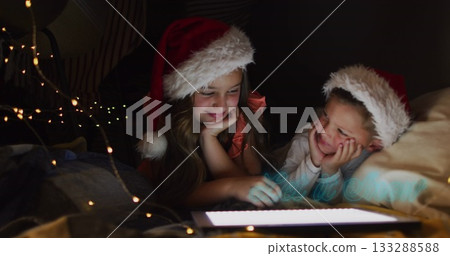 Lying siblings using glowing tablet in bedroom at home, with Santa hats and string lights Lying siblings using glowing tablet in bedroom at home, with Santa hats and string lights 133288588