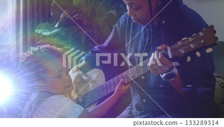 Mother in blue shirt guiding toddler strumming acoustic guitar near living room window, with plant Mother in blue shirt guiding toddler strumming acoustic guitar near living room window, with plant 133289314