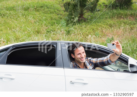 Man leaning out open window of white sedan taking smartphone selfie by grassy hillside, copy space 133290597