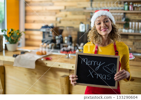 Smiling female barista wearing Santa hat holding Merry Xmas chalkboard at cafe counter, copy space 133290604