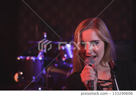 Woman holding microphone while singing on small stage beside drum kit under red lights, copy space 133290606
