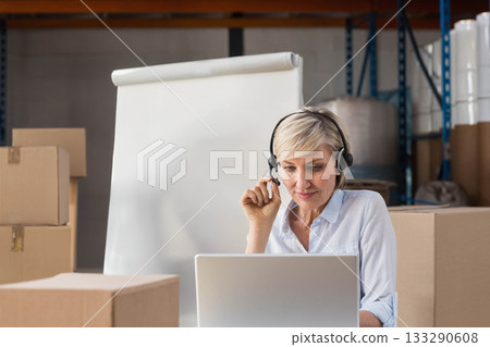 Mature adult woman sitting on cardboard boxes using headset and laptop near flip chart in warehouse Mature adult woman sitting on cardboard boxes using headset and laptop near flip chart in warehouse 133290608